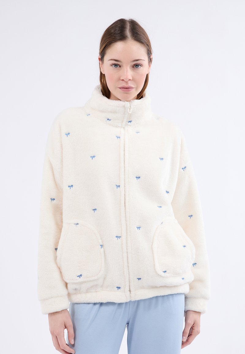 Puszysta kremowa kurtka z niebieskimi haftowanymi wzorami w kształcie kokardy, z wysokim kołnierzem, zamek z przodu i dwoma bocznymi kieszeniami. Miękka w dotyku, luźny krój.