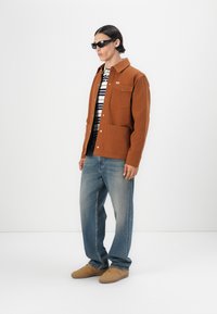 Bruine, knoopsluiting jas met twee voorzakken, over een gestreept shirt. Gecombineerd met lichtblauwe jeans en tan sneakers. Model draagt zwarte zonnebril.