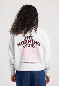 Lichtgrijze sweatshirt met een groot grafisch ontwerp op de achterkant dat "THE MORNING CLUB" in bordeauxrood weergeeft, met daaronder een ontbijtillustratie.