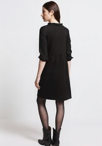 Schwarzes Cordkleid mit gerafftem Kragen, dreiviertellangen Ärmeln und einem taillierten Oberteil, das in einen ausgestellten Rock übergeht. Kombiniert mit schwarzen Stiefeletten.