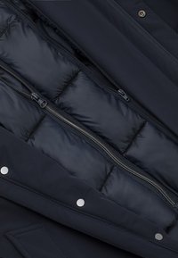 Navy puchowa kurtka od wewnątrz z pikowanym wypełnieniem, z suwakiem i metalowymi zatrzaskami. Materiały wydają się gładkie i ocieplające.