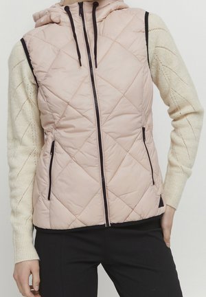 Žena má na sobě béžovou prošívanou vesta bez rukávů s kapsami, přes svetr krémové barvy a černé kalhoty, stojí s rukama u kapes.