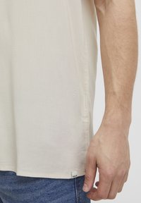 Beige kortermet skjorte laget av lett stoff. Kanten har en liten merkeetikett. Skjorten har en avslappet passform med en jevn tekstur.