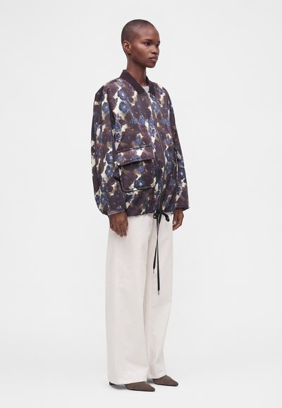 Mannequin portant une veste bomber ample fleurie foncée avec de grandes poches avant et un pantalon blanc à jambes larges sur un fond blanc uni.