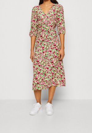 Kobieta ubrana w sukienkę do kolan w kwiaty w kolorach różowym, czerwonym i zielonym, z dekoltem w serek, rękawami do łokci oraz białymi trampkami.