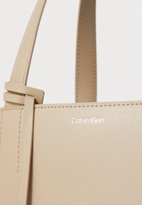 Bež torba od umjetne kože sa strukturiranim dizajnom, koja ima uski remen i utisnutu "Calvin Klein" oznaku u bijeloj boji na prednjoj strani.