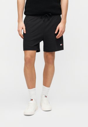 Bărbat purtând pantaloni scurți negri cu șnur, șosete albe și adidași albi cu logo-uri de brand pe pantaloni și pe încălțăminte.