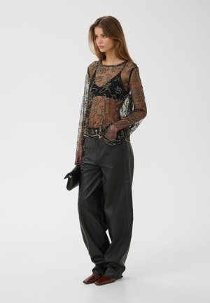 Femme portant un haut noir transparent brodé de fleurs sur un soutien-gorge noir, un pantalon en cuir noir à jambes larges, des chaussures marron, et tenant une pochette noire.