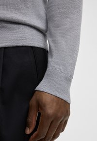 Grauer Pullover mit geripptem Bündchen, glatter Textur und figurbetontem Design, kombiniert mit dunklen Hosen. Nahaufnahme des Arms und der Hand.