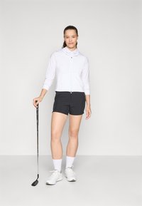 Bijela majica s dugim rukavima na zip, crne šortse i bijele tenisice. Golf klub ležerno drži uz tijelo, stojeći na neutralnoj pozadini.