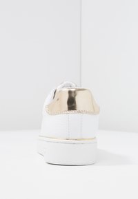 Baskets blanches avec un accent brillant doré au talon, une tige texturée et une semelle en caoutchouc lisse, avec des détails embossés subtils sur le côté.