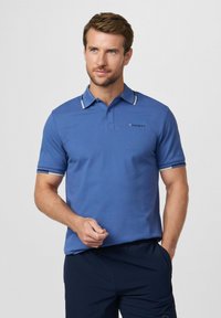 Homme portant un polo à manches courtes bleu avec le logo "Hackett" sur la poitrine, pantalon bleu marine, regardant légèrement sur le côté avec une main dans la poche.