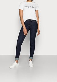 Dunkelblaue Skinny Jeans mit aufgerollten Säumen, kombiniert mit einem weißen T-Shirt mit schwarzem, geschwungenem Logo-Stickerei und weißen Turnschuhen.