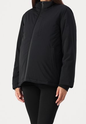 Vrouw draagt een zwarte, asymmetrisch ritsende winterjas met borst- en zijzakken, gecombineerd met een zwarte broek tegen een effen achtergrond.