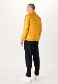 Gele gewatteerde jas met verticale gestikte patroon, hoge kraag en ribbels bij de manchetten, gecombineerd met zwarte broeken en witte sneakers.