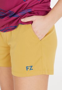 Person klädd i senapsgula shorts med "FZ"-logotyp och en magenta t-shirt med blå mönster, hand vilande i shortsfickan.