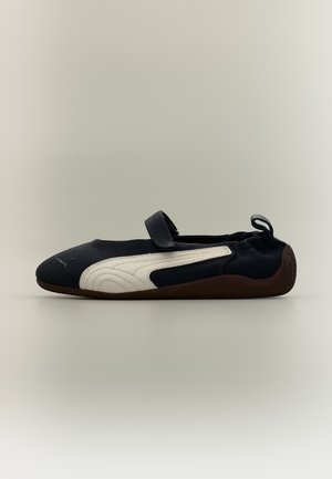 Une chaussure slip-on noire et blanche avec semelle marron, dos élastique et bande Velcro sur le dessus, vue de profil latéral.