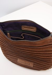 Brązowa torba z zamszu o karbowanej fakturze, z zamknięciem na zamek, wewnętrzną kieszenią i fioletowym wykończeniem, widoczna embosowana etykieta z logo w pobliżu zamka.