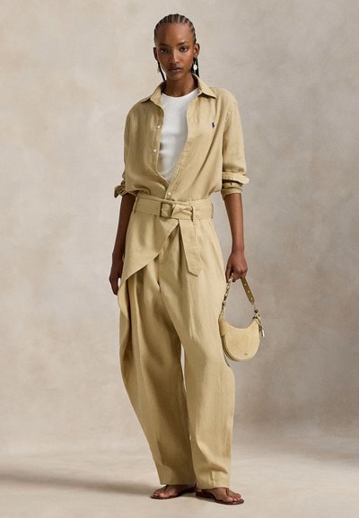Μπεζ λινό πουκάμισο με μανίκια γυρισμένα, πάνω από λευκό ριμπ αμάνικο τοπ, συνδυασμένο με χαλαρό, ψηλόμεσο μπεζ παντελόνι και μικρή μπεζ τσάντα χειρός.