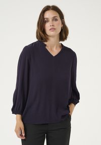 Navy blue blouse with a V-neckline, gathered details at shoulders, and long, puffed sleeves. Smooth texture, paired with black pants.