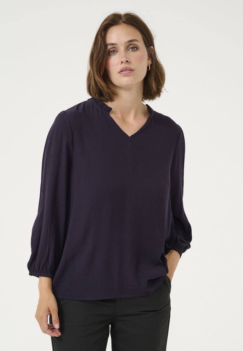 Navy blue blouse with a V-neckline, gathered details at shoulders, and long, puffed sleeves. Smooth texture, paired with black pants.