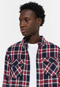 Geruite flanellen shirt in marineblauw, rood en wit. Heeft een button-down kraag, twee borstzakken en een gestructureerde stof. Witte T-shirt eronder.