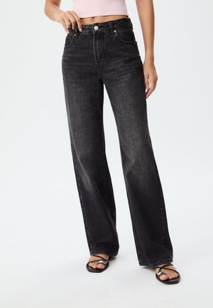 High-waisted black denim jeans with a wide-leg fit, featuring five pockets and subtle fading, paired with black flip-flops.