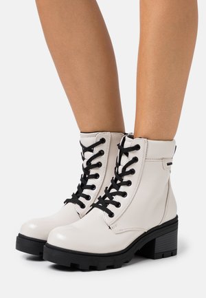 Bílé kožené kotníkové boty s černými šněrovadly, robustním podpatkem a texturovaným povrchem. Mají kontrastní černou podešev a střední střih.
