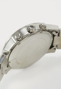 Reloj de acero inoxidable plateado con caja redonda, tres botones de presión y una pulsera de metal texturizada, que presenta un acabado suave y pulido.