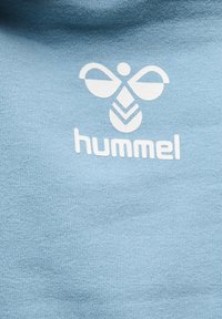 Hellblaues Baumwoll-T-Shirt mit weicher Textur, das ein weißes Logo mit stilisierten Flügeln und dem Markennamen "hummel" darunter zeigt.
