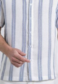 Chemise blanche à manches courtes avec des rayures verticales bleues. Tissu texturé et patte de boutonnage avec des boutons ronds et transparents.