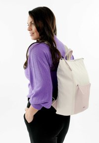 Zaino in crema e rosa chiaro, realizzato in materiale liscia, con una tasca anteriore con zip e spallacci regolabili. Modella con un maglione viola.