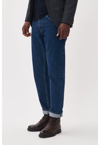 Jeans de mezclilla azul oscuro con un corte relajado, con los tobillos doblados, combinados con botas de cuero marrón con cordones y un blazer gris. Textura suave y tejido resistente.