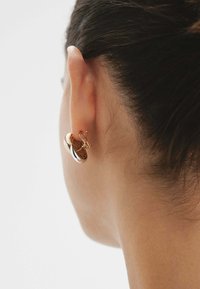 Pendientes en tono dorado con un diseño circular, que presentan una textura tejida y acentos suaves, mostrados en la oreja de un modelo desde un lado.
