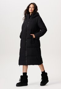 Zwarte lange pufferjas met een capuchon, frontale ritssluiting en zijzakken. Kenmerkt zich door gestikte panelen en een ontspannen pasvorm. Gecombineerd met zwarte fleece laarzen.