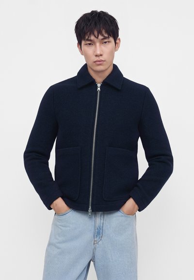 Jeune homme aux cheveux noirs portant une veste zippée bleu marine foncé et un jean bleu clair, debout les mains dans les poches devant un fond uni.