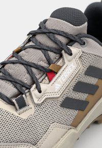 Beżowe siatkowe buty sportowe z ciemnoszarymi sznurówkami, gumowymi akcentami i logo 'Terrex'. Posiadają zaokrąglony czubek i fakturowaną powierzchnię.