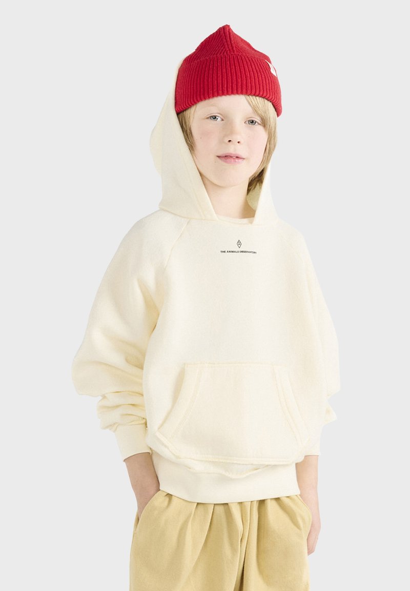 Felpa color crema realizzata in morbido tessuto, con tasca frontale, grande cappuccio e logo del marchio stampato sul petto. Abbinata a pantaloni beige.
