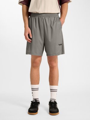 Mężczyzna w szarych sportowych szortach, białe skarpetki z czarnymi paskami i czarne trampki z gumowymi podeszwami, stoi z rękami w kieszeniach.