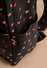 Juodos medžiagos kuprinė su raudonų, rožinių ir oranžinių žvaigždžių raštais, turinti šoninę kišenę ir auksinės spalvos metalinę žiedinę detalę.