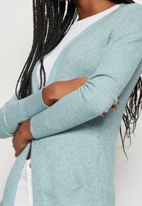 Jasnoniebieski sweter z dzianiny z długimi rękawami, otwarty z przodu i z prążkowanymi mankietami. Miękka faktura i luźny krój. Brak widocznych zapięć lub wzorów.