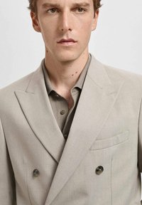 Blazer doppio petto color beige chiaro con rever a punta, due bottoni e taschino sul petto, abbinato a una camicia button-up color taupe.
