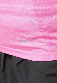 Vaaleanpunainen urheilupaita, jossa on verkkopaneeli ja pieni harmaa Nike-swoosh-logo, mustien housujen päällä.