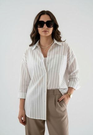 Femme portant une chemise blanche oversize à rayures verticales noires, un pantalon beige, des lunettes de soleil noires, un collier de perles et une montre en or, posant avec une main dans la poche.