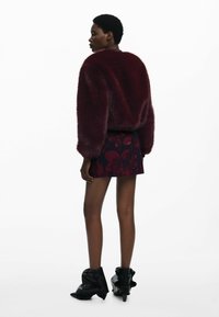 Chaqueta de piel sintética burdeos con una silueta redondeada, llevada sobre una falda corta con estampado floral, combinada con botas de tobillo negras y de punta.