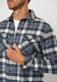 Tamsiai mėlynos, baltos ir oranžinės spalvų languota marškiniai. Su dviem užsegamais krūtinės kišenėmis, apykakle ir ilgomis rankovėmis su rankogalių detalėmis. Pagamintas iš medvilnės.