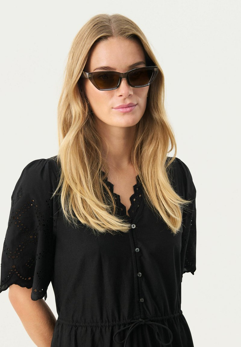 Des lunettes de soleil en œil de chat avec des verres teintés foncés et une monture transparente. Le modèle porte une robe noire à boutons avec des broderies sur les manches.