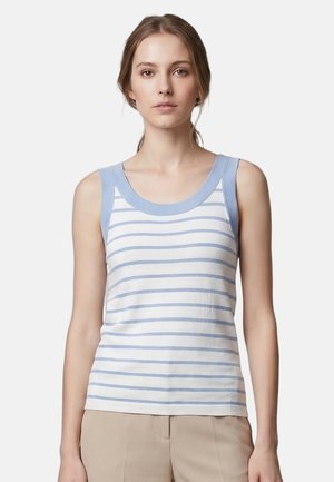 Femme portant un haut blanc sans manches avec des rayures horizontales bleu clair et un pantalon beige, debout devant un fond blanc uni.