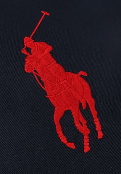 Κόκκινη κεντημένη φιγούρα παίκτη πόλο που ιππεύει άλογο, κρατώντας υψωμένη ματσέτα, σε σκοτεινό φόντο υφάσματος.