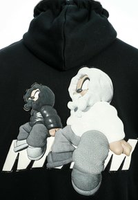 Dos personajes de estilo caricaturesco con trajes con capucha en blanco y negro sentados sobre un gran texto blanco en una sudadera negra.